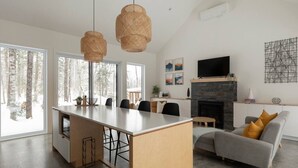 A modern living room with a fireplace, a dining area, and large windows with a snowy view. - Woodland Cocoon | Fireplace • 15min Tremblant (Les Laurentides Regional County Municipality)