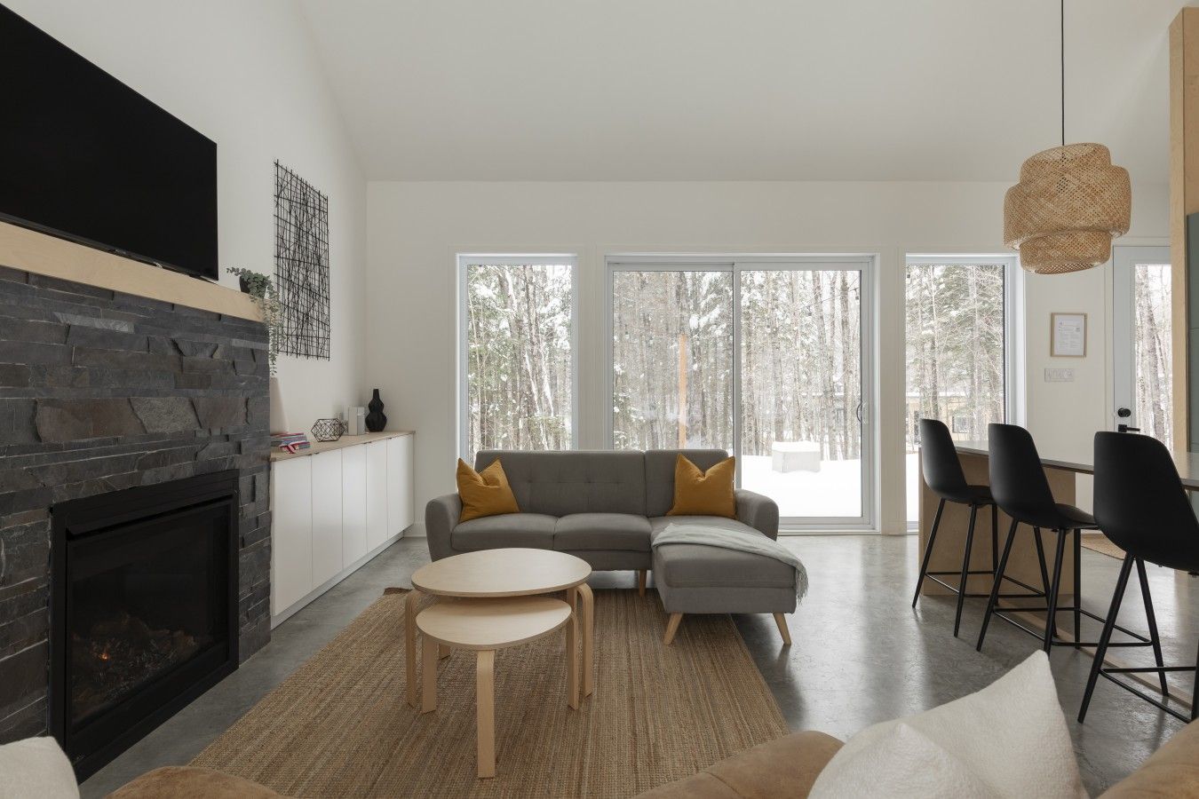 Uma sala de estar moderna com lareira de pedra, sofá modular cinza com almofadas amarelas, mesa de centro redonda de madeira e uma área de jantar com cadeiras pretas.