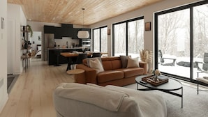 A modern living room with a wooden ceiling, large windows, a sofa, a coffee table, and a kitchen in the background.