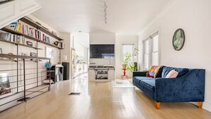 A spacious living area with a blue sofa, a bookshelf, and a kitchen in the background. - Spacious, artful 4-bedroom house in Brooklyn with lush backyard, parking, A/C (Brooklyn)