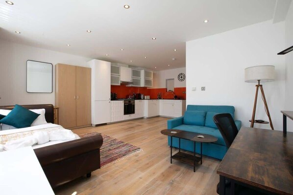 A modern hotel room with a bed, a sofa, a small table, a lamp, and a kitchenette in the background.