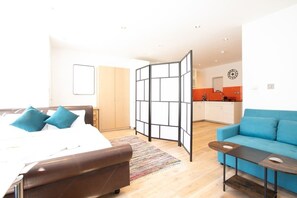 A modern hotel room with a bed, a sofa, a small table, a kitchen area, and a partition.