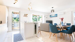 A modern kitchen with a built-in oven, a dining area with blue chairs, and a large window offering a view of greenery and buildings. - 4* apartment | swimming pool | parking | wifi (Saint Raphaël)
