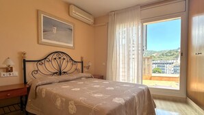 Un dormitorio con cama, mesita de noche, lámpara, un cuadro y ventana con vistas.