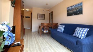 Una sala de estar con un sofá azul, una mesa de comedor de madera y sillas. Hay un televisor sobre un mueble de madera, una planta y un cuadro en la pared.