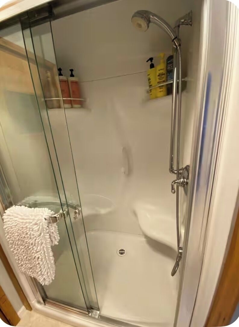 Et brusebad med glasvægge, en håndbruser og et håndtag med vandhane, samt en glasdør.
