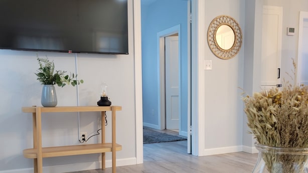 A modern living room with a flat-screen TV mounted on the wall, a wooden TV stand with a vase of flowers, a remote control on a wooden table, and a mirror on the wall.