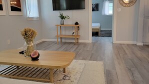 A modern living room with a wooden table, a flat-screen TV mounted on the wall, a small wooden shelf, and a potted plant.