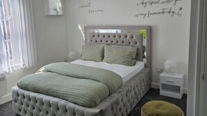 A neatly arranged bedroom with a tufted headboard, a bed with green bedding, a bedside table with a lamp, and a small woven stool. - 1 Bed - Sleeps 4 - City Centre (Wolverhampton)