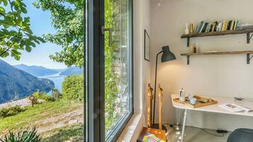 Une pièce dotée d’une grande fenêtre donnant sur un paysage montagneux. On y trouve un bureau avec une lampe, des étagères à livres, ainsi qu’un plateau contenant des livres et une tasse.
