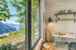 Una habitación con un amplio ventanal que da a un paisaje montañoso. Hay un escritorio con una lámpara, estanterías y una bandeja con libros y una taza.