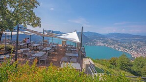 An outdoor dining area with tables and chairs, overlooking a coastal city and a lake.