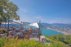 An outdoor dining area with tables and chairs, overlooking a coastal city and a lake.