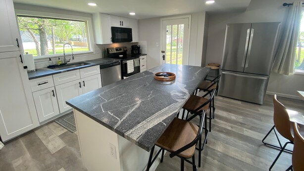 A modern kitchen with white cabinets, a black countertop, stainless steel appliances, and a large island with bar stools. - Monkey Island Escape sleeps 8, surrounded by nature with views of Grand Lake. (Monkey Island)