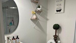 A bathroom with a white toilet, a blue vanity, a round mirror, a shower enclosure, and a wall-mounted shelf with toiletries. - Home away from home with WiFi and AC in delightful Parkville (Parkville)