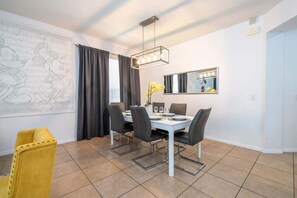 Uma sala de jantar com uma mesa branca, cadeiras pretas, uma cadeira amarela, um lustre, um espelho com iluminação e uma parede com o desenho do Mickey Mouse.