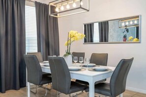 A dining area with a white table, black chairs, a vase with yellow flowers, and a wall-mounted mirror with lights.