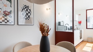 A modern dining area with a round wooden table, a dark vase with dried plants, and two chairs. There are framed artworks on the wall, a kitchen area visible through an open door, and a red fire safety sign on the wall.