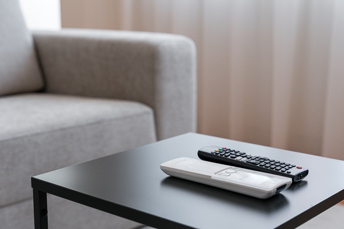 A remote control and a white device on a black table in a room with a grey sofa.