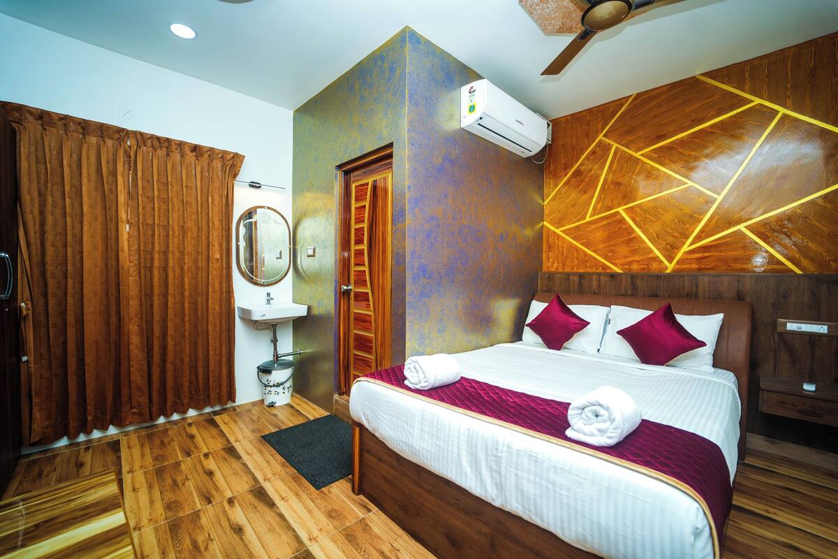 A hotel room with a wooden floor, a bed with white bedding and red pillows, a wall-mounted air conditioner, and a wooden headboard.