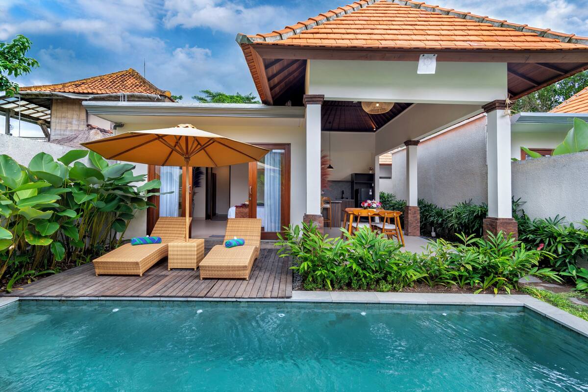 A pool area with lounge chairs and an umbrella, surrounded by greenery and a covered patio.