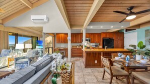A spacious living area with a sofa, dining table, and chairs, a kitchen with wooden cabinets, and a ceiling fan.