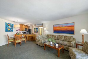 A living room with a sofa, coffee table, and dining area with chairs. There is a kitchen in the background.