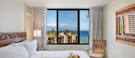 Quarto de hotel com cama, cadeira, janela com vista para o oceano e palmeiras, e um espelho decorativo.