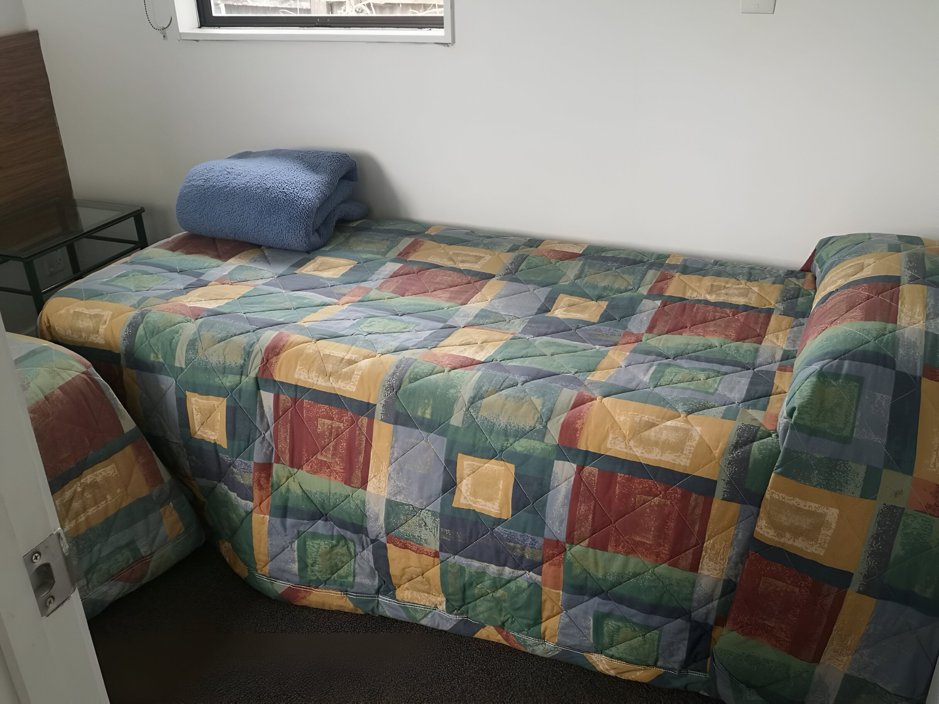 Una habitació petita i acollidora amb un estovat de llit colorit, una manta blava i una finestra amb vista a l'exterior.