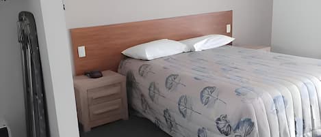 Habitación de hotel con cama, cabecera de madera, mesita de noche con lámpara y armario.