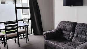 Ein Zimmer mit einer Couch, einem an der Wand montierten Fernseher, einem Esstisch mit Stühlen und einem Fenster mit Vorhängen.