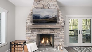 Ein Kamin aus Stein mit einem Fernseher darüber, ein Feuer brennt und ein Blick auf Berge durch das Fenster.