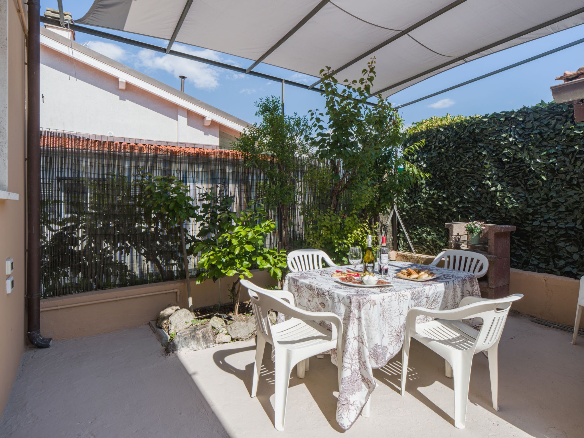 A patio with a table set for two, surrounded by white chairs, under a shaded area with a view of a garden and a stone wall.