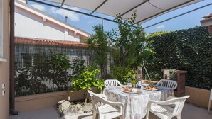 Un patio con una mesa lista para dos personas, rodeado de sillas blancas, bajo una zona sombreada con vistas a un jardín y un muro de piedra.