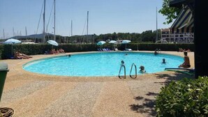 A swimming pool with lounge chairs and umbrellas, surrounded by greenery and a building in the background.