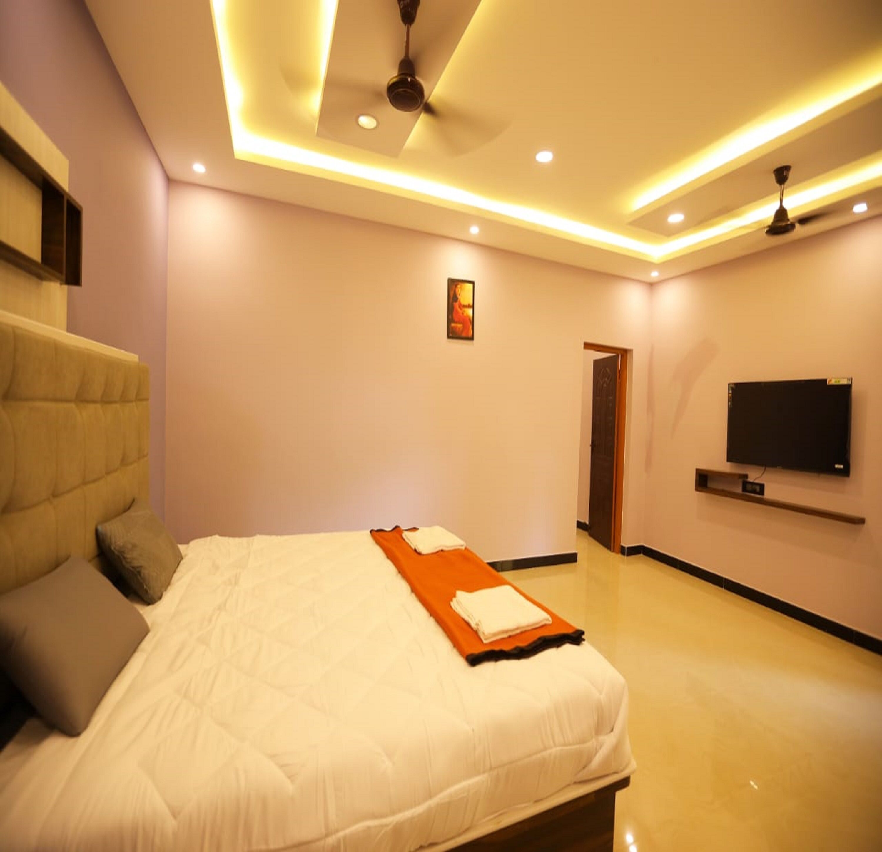 A modern bedroom with a bed, pillows, a ceiling fan, recessed lighting, a flat-screen TV, and a door.