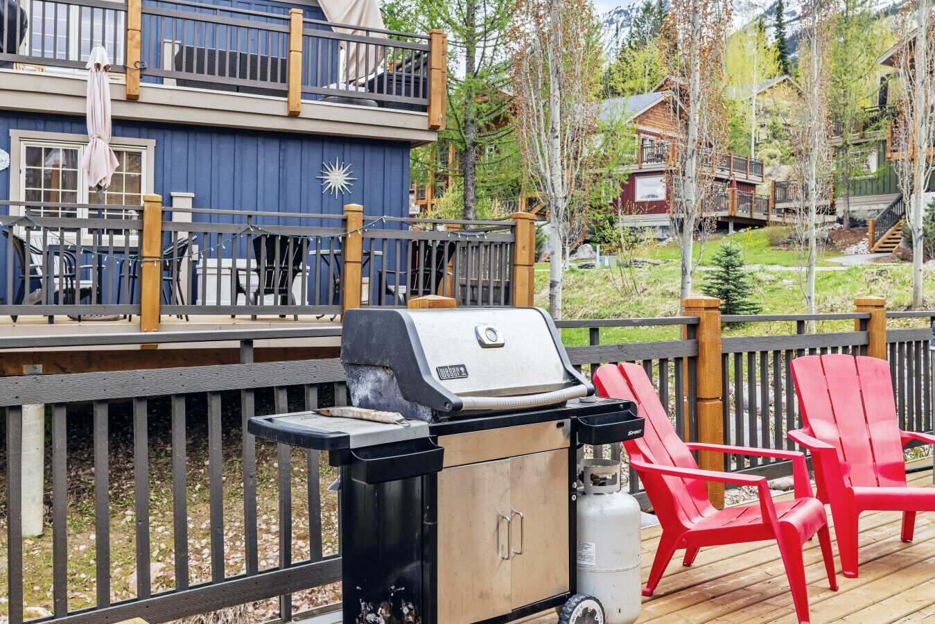 A deck with a barbecue grill, red Adirondack chairs, and a view of a cabin in the background.