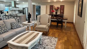 Una sala de estar moderna con un sofá, una mesa de centro y elementos decorativos. Se observa un comedor con una mesa y sillas, y una cocina al fondo.