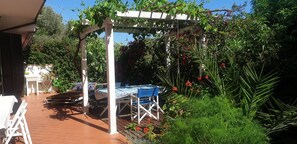 A covered outdoor seating area with a table and chairs, surrounded by lush greenery and flowering plants.
