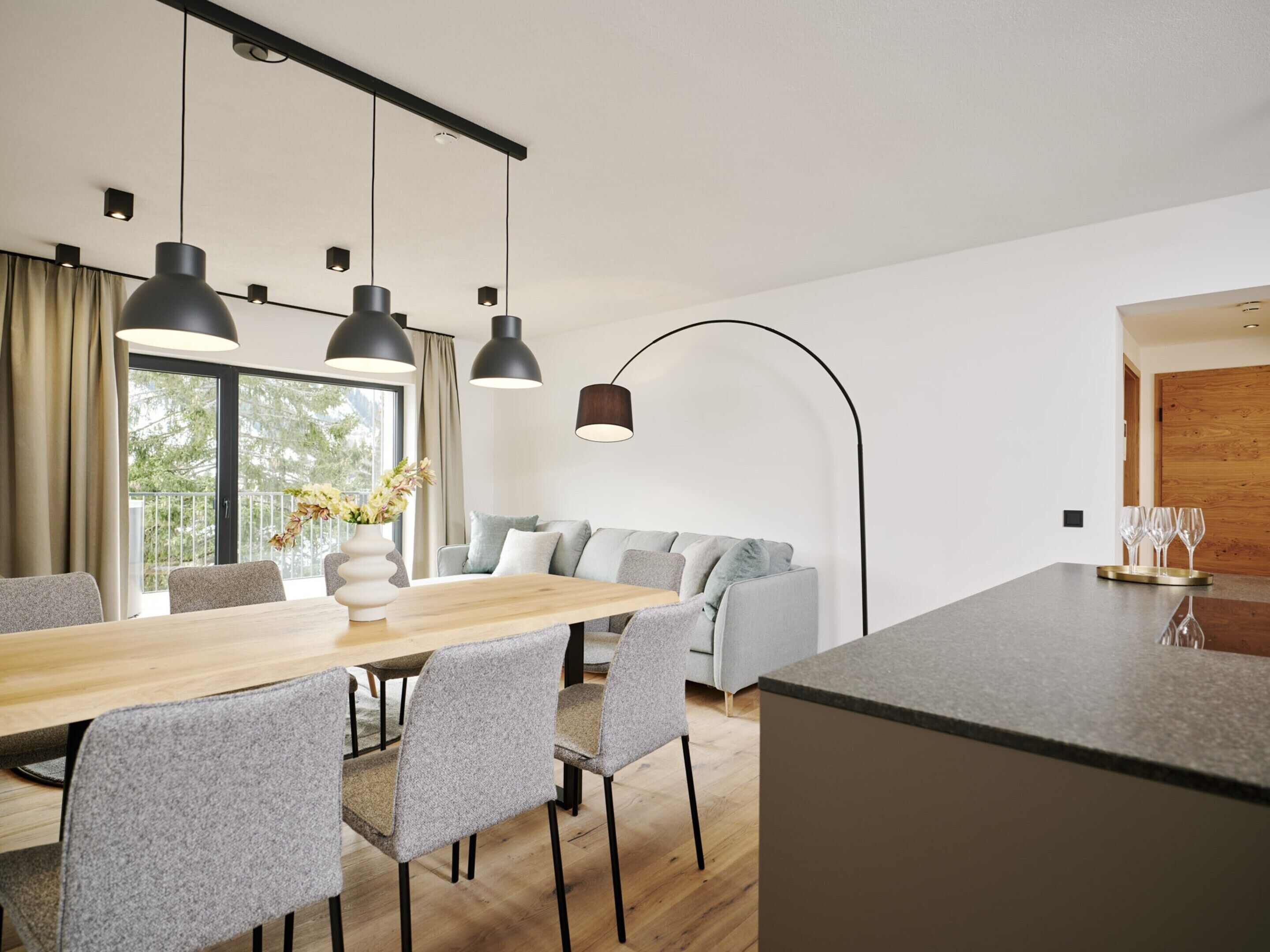 A modern kitchen with a wooden dining table, grey upholstered chairs, and pendant lighting.