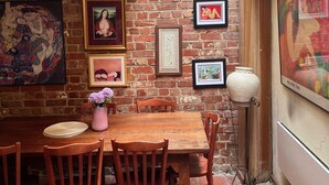 A dining area with a brick wall, wooden table, and chairs. There are framed pictures on the wall and a vase with flowers on the table.