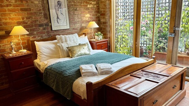 A bedroom with a wooden bed, two bedside tables with lamps, a wooden dresser, and a view of a garden through a glass door.