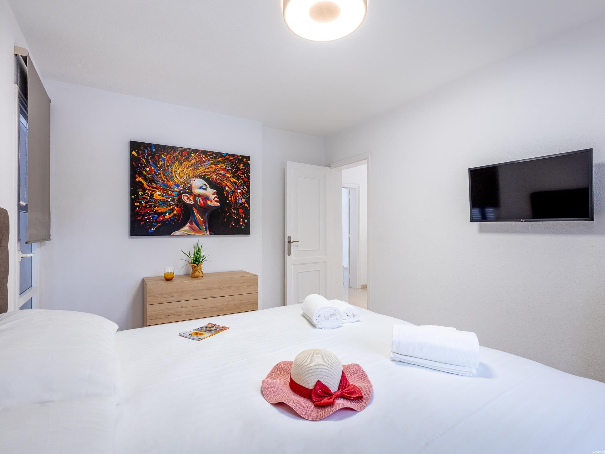 A hotel room with a bed, a painting on the wall, a TV mounted on the wall, and a wooden dresser.