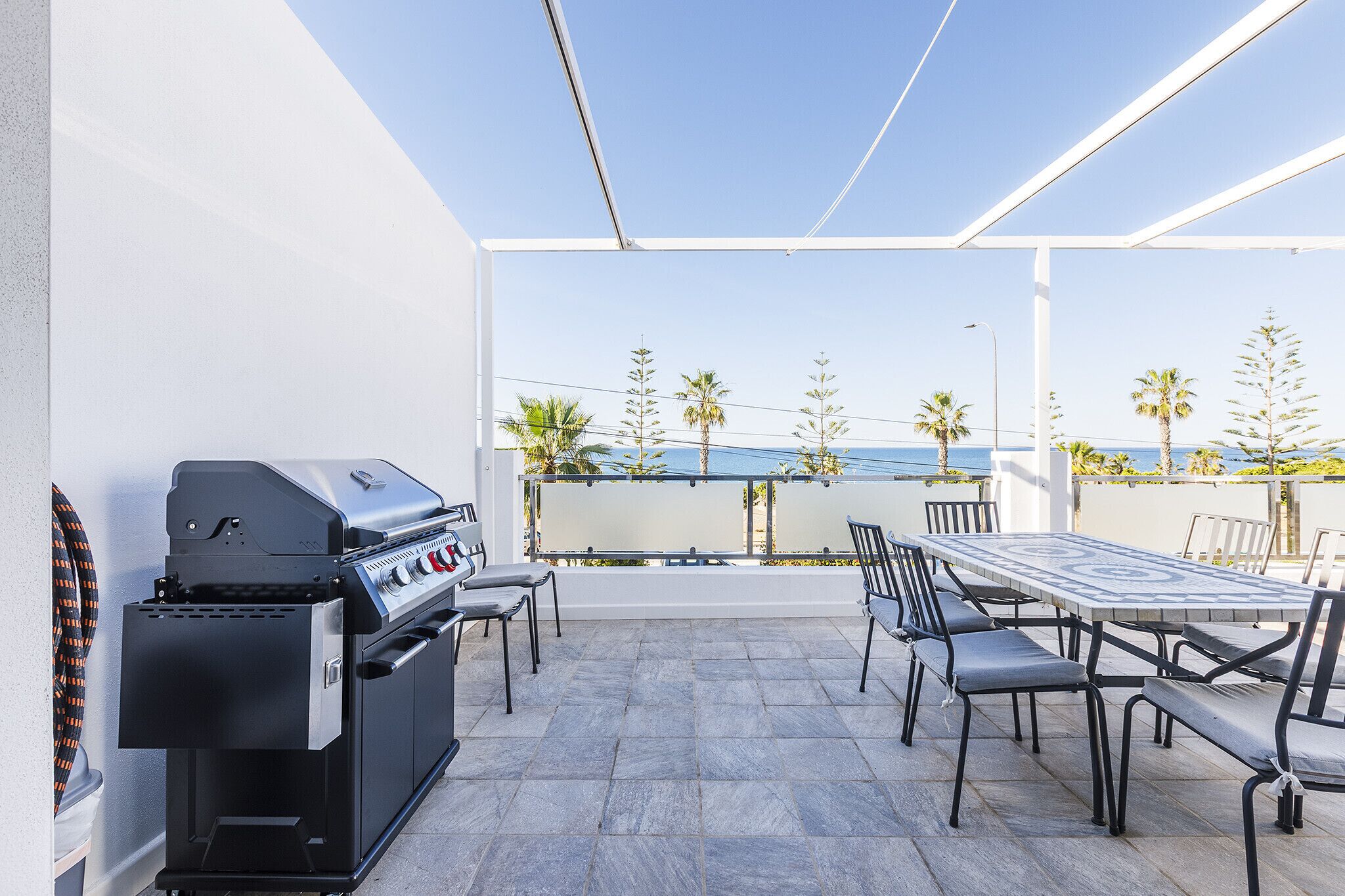A patio with a grill, outdoor table, and chairs, overlooking a coastal area with palm trees.
