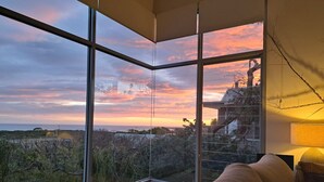 Quarto com grandes janelas que oferecem vista para um pôr do sol sobre a paisagem costeira.