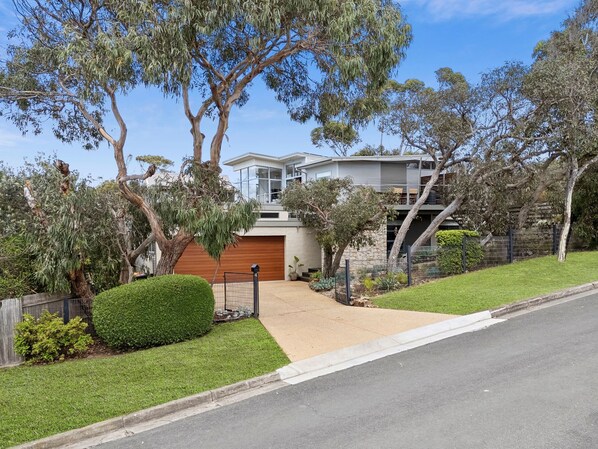 Une maison moderne à deux étages avec un garage, entourée de pelouses et d’arbres bien entretenus.