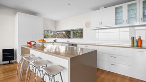 Uma cozinha moderna com armários brancos, uma ilha central com banquetas e uma janela com vista para uma árvore.