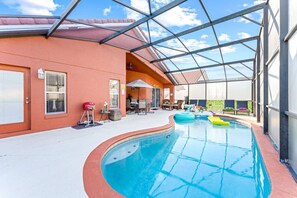 Une piscine intérieure dotée d’un toit en verre, entourée d’un patio aménagé avec des espaces de détente en plein air et un barbecue.
