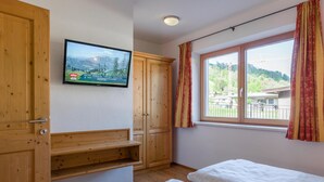 Et værelse med møbler af træ, et fjernsyn der viser et bjerglandskab, et vindue med gardiner og udsigt til grøn natur udenfor.