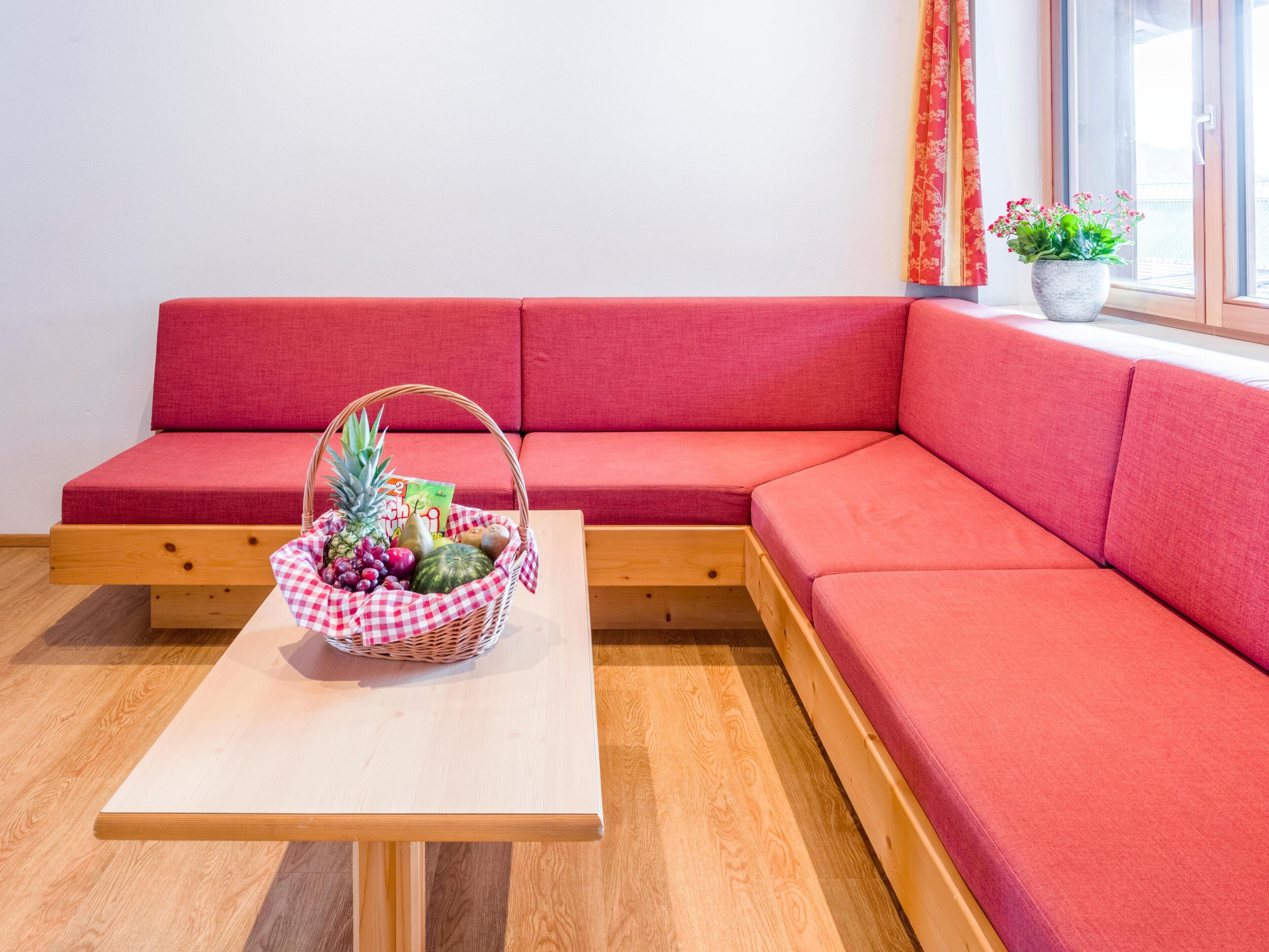 Ein rotes Sofa mit einem Holztisch davor, ein Korbwäscher mit Früchten und ein Fenster mit Vorhängen.